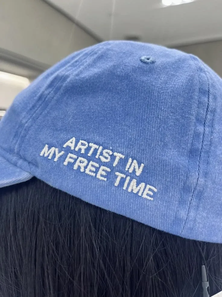 White block lettering reading artist in my free time on the back of a blue cap